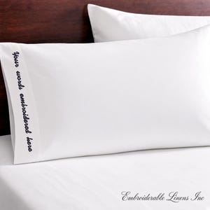 May include: A crisp white pillowcase featuring a vertical band on the left side with the black embroidered text "Your words embroidered here" in a cursive script font. The pillowcase is smooth and clean, presented against a dark wooden headboard.