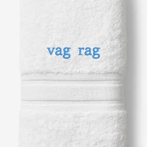 May include: White folded towel with the text "vag rag" in blue. The towel has a textured surface and a decorative band. The words "Embroiderable Linens Inc." are printed at the bottom.