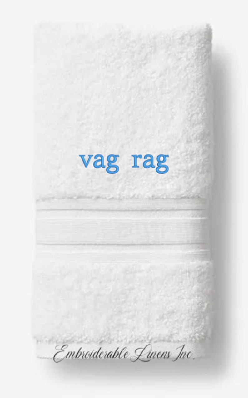 May include: White folded towel with the text "vag rag" in blue. The towel has a textured surface and a decorative band. The words "Embroiderable Linens Inc." are printed at the bottom.