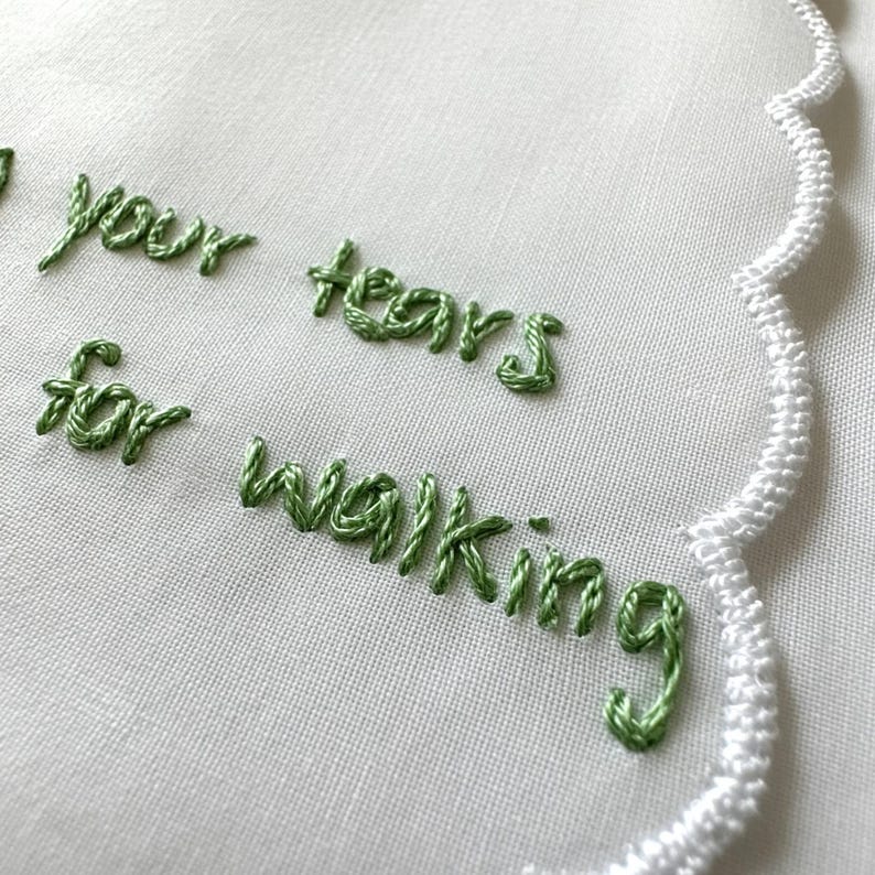 K&ouml;nnte beinhalten: Ein wei&szlig;es Stofftaschentuch mit gewelltem Rand und gr&uuml;ner Stickerei mit dem Text your tears for walking. Die Stickerei weist einen erhabenen, strukturierten Stichstil auf einer glatten, schlicht gewebten wei&szlig;en Textiloberfl&auml;che auf.