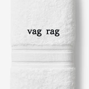 May include: White, folded towel with the text "vag rag" in black. The towel has a textured surface and a decorative band near the top. The words "Embroiderable Linens Inc." are printed at the bottom.