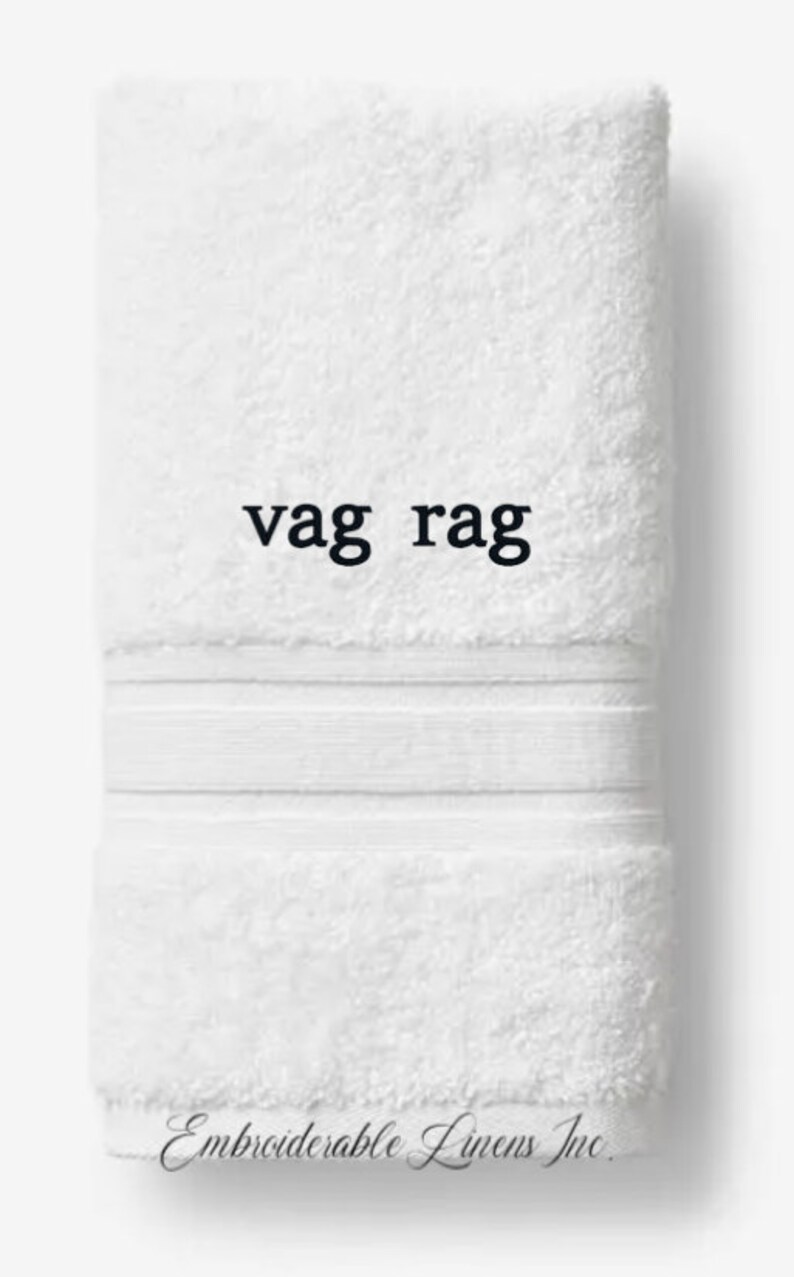 May include: White, folded towel with the text "vag rag" in black. The towel has a textured surface and a decorative band near the top. The words "Embroiderable Linens Inc." are printed at the bottom.
