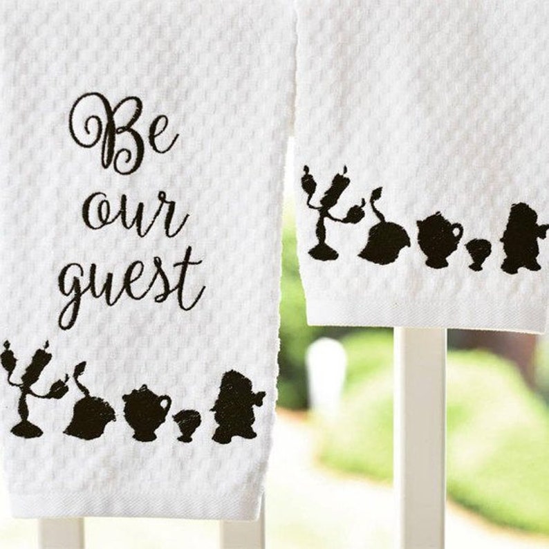 May include: Two white towels with black embroidery. One towel says "Be our guest" and the other has a silhouette of characters from Beauty and the Beast.