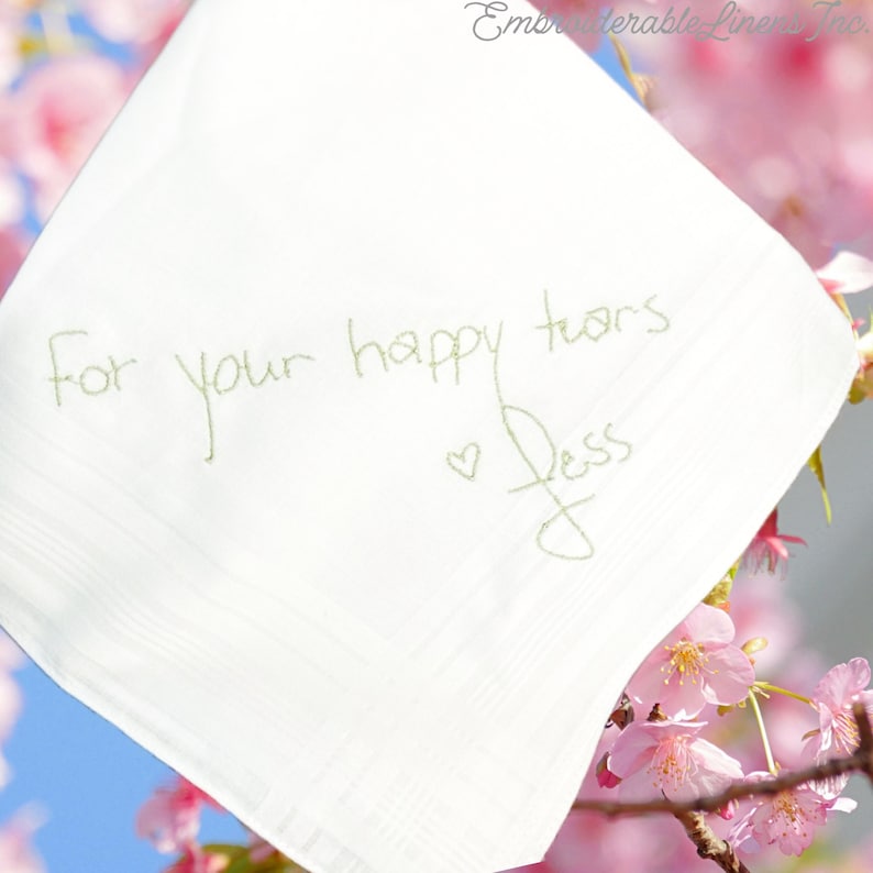 K&ouml;nnte beinhalten: Ein wei&szlig;es quadratisches Taschentuch mit hellgr&uuml;n gesticktem Text, der For your happy tears lautet, gefolgt von einem kleinen Herz und dem Namen Jess. Der Stoff hat ein dezentes gewebtes Randmuster und ist vor einem Blumenhintergrund.