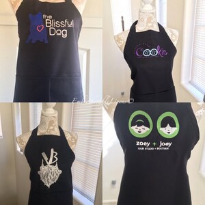 May include: Four black aprons with different embroidered designs. The first apron has a blue dog with a heart and the text "The Blissful Dog". The second apron has the text "Enough to Eat a Cookie". The third apron has a white beard with the letters "VB". The fourth apron has two cartoon girls with the text "Zoey + Joey Hair Studio + Boutique".