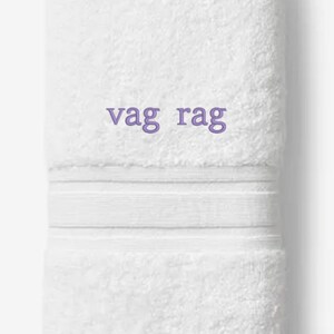 May include: White, folded towel with the embroidered text "vag rag" in purple. The towel has a textured surface and a decorative band. The words "Embroiderable Linens Inc." are printed at the bottom.