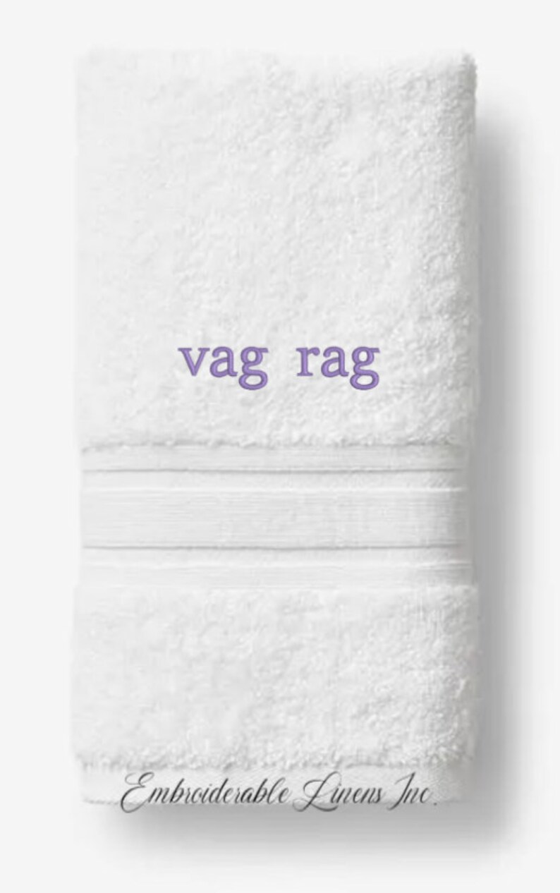 May include: White, folded towel with the embroidered text "vag rag" in purple. The towel has a textured surface and a decorative band. The words "Embroiderable Linens Inc." are printed at the bottom.