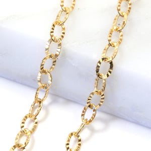 May include: Close-up of two strands of a gold-coloured chain. The chain links are oval-shaped with a textured, patterned design. The chains are displayed against a white marble surface.
