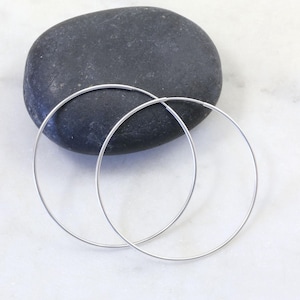 May include: A pair of thin, circular silver hoop earrings with a smooth, polished finish. The earrings are positioned in front of a single, smooth, dark gray oval stone, showcasing their simple, open-loop design and metallic silver color.