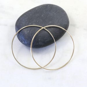 Puede incluir: Un par de aros circulares finos de oro con un acabado liso y pulido. Los pendientes son sencillos, ligeros y presentan un diseño de alambre continuo que forma un círculo perfecto, mostrando una estética metálica clásica y discreta.