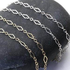 May include: Three silver and gold chains with a diamond-shaped link pattern are displayed on a dark grey stone. The chains are arranged in a layered fashion, with the gold chain on top.