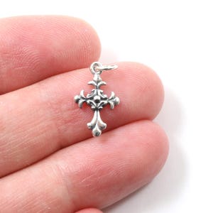 Peut inclure: Un petit pendentif en forme de croix argentée ornée d'un motif détaillé. La croix présente un style fleur-de-lis aux extrémités de chaque bras et une boucle en haut pour l'attacher à une chaîne. Le pendentif est tenu dans une main sur fond blanc.