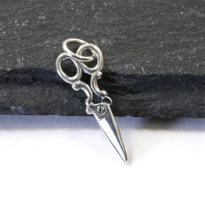 May include: A small, silver-colored pendant in the shape of a pair of scissors. The scissors have a detailed design with looped handles and a pointed blade. The pendant is likely made of metal and is displayed on a dark, textured surface.
