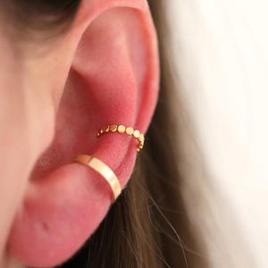 May include: Close-up of an ear adorned with two gold ear cuffs. One cuff is a simple band, while the other features a series of small, connected circles. The jewelry is set against a soft, neutral background, highlighting the delicate design.