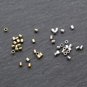 May include: A collection of small, cylindrical beads in gold and silver tones scattered on a dark gray surface. The beads appear to be made of metal and are evenly cut, suggesting they are for jewelry making or crafting.