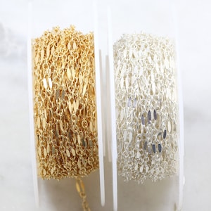 Puede incluir: Dos carretes de cadena, uno dorado y otro plateado, con un pequeño tramo de cada cadena colgando. La cadena está hecha de pequeños eslabones planos de forma ovalada.