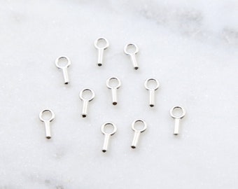 May include: A collection of small, silver-colored key charms scattered on a white marble surface. Each charm features a keyhole-shaped top and a cylindrical stem. The charms are evenly spaced, creating a visually appealing arrangement.