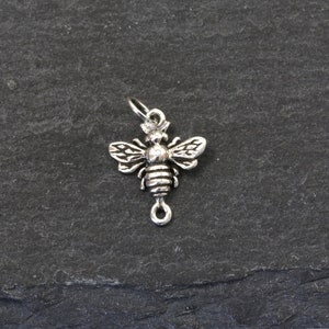 May include: A small, silver-colored bee charm with detailed wings, body segments, and a loop for attachment. The charm is set against a dark, textured background, highlighting its intricate design. Ideal for jewelry making or as a pendant.