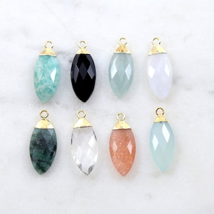 May include: Eight gemstone pendants with gold-colored caps. The gemstones are in various colors, including teal, black, clear, and peach. The pendants are teardrop-shaped and faceted, displayed on a white marble surface.