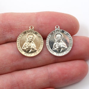 May include: Two small, round, metal charms with a gold and silver finish. Both charms feature an image of Jesus with the words "Sacred Heart of Jesus Have Mercy on Us".