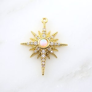 May include: Gold-coloured starburst pendant featuring a central opal-like stone and clear crystals. The pendant has multiple pointed rays and a small loop for hanging. The jewellery piece is set against a white background.