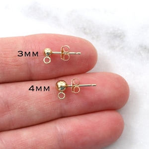 May include: Two gold earring findings, one 3mm and one 4mm in size, with a loop on the end. The findings are shown on a hand.