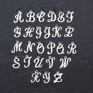 May include: A set of 26 silver-toned metal alphabet letter charms in a cursive font. The letters are arranged in alphabetical order on a black background.