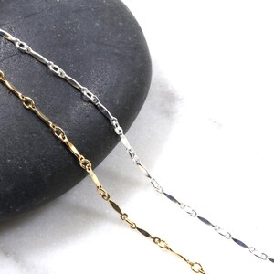 May include: Two delicate chains, one silver and one gold, lay on a dark gray stone background. The chains are made of small, elongated links.