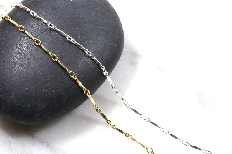 May include: Two delicate chains, one silver and one gold, lay on a dark gray stone background. The chains are made of small, elongated links.