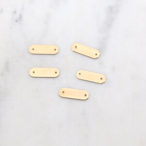 May include: Five gold-colored metal rectangular tags with two holes each. The tags are on a white marble surface.