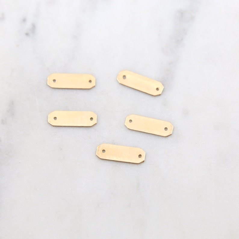 May include: Five gold-colored metal rectangular tags with two holes each. The tags are on a white marble surface.