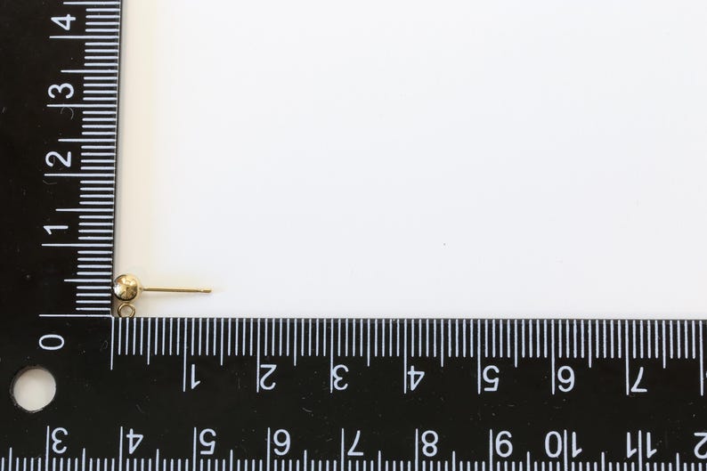 May include: A gold-toned earring finding with a small ball at the top and a straight post. The finding is measured against a ruler.