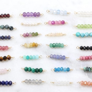 Peut inclure: Une collection de connecteurs de perles de pierres précieuses de différentes couleurs, dont le violet, le rose, le vert et le bleu. Chaque connecteur présente une rangée de perles à facettes enfilées sur du fil doré avec des boucles à chaque extrémité. Les perles sont disposées sur une surface blanche.