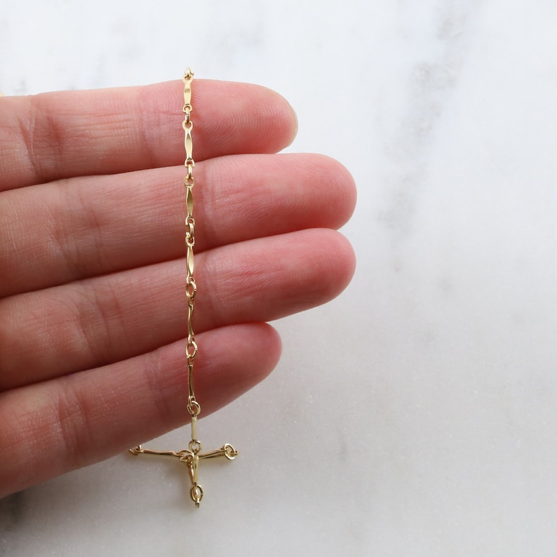 May include: A gold chain necklace with a delicate, elongated link design. The chain is held in a hand, and the end of the chain has a small, simple cross pendant.