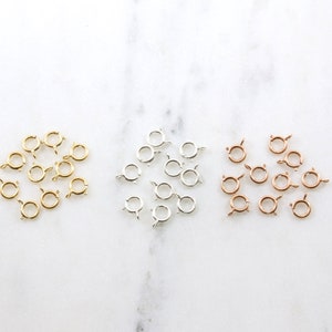 May include: An assortment of jewelry clasps in gold, silver, and rose gold tones. The clasps are small, circular, and have a spring-loaded mechanism for secure fastening. They are arranged on a white marble surface.