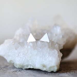 May include: A pair of silver triangle stud earrings on a white crystal surface.