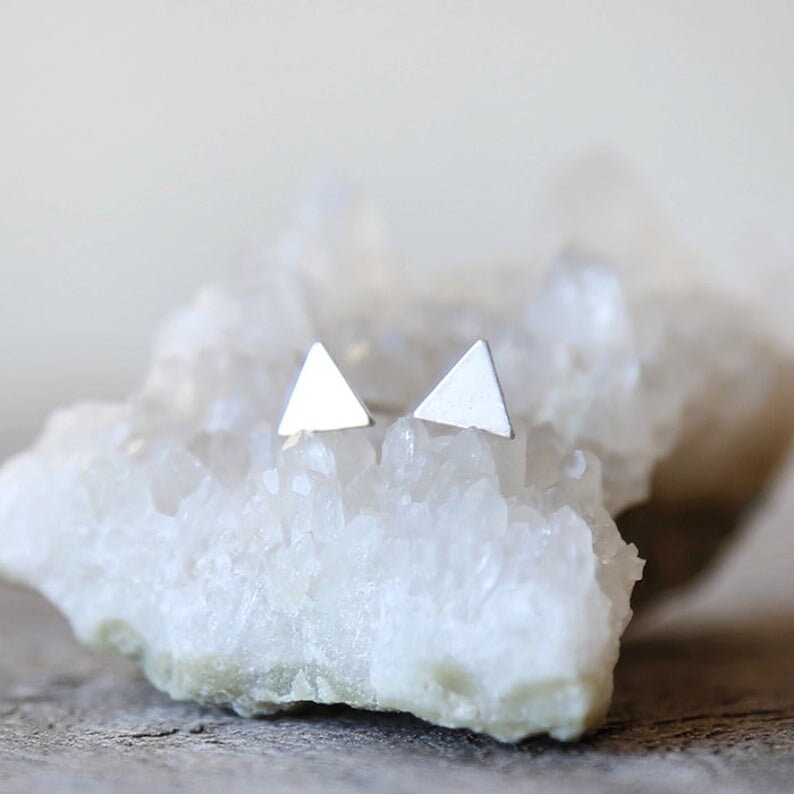 May include: A pair of silver triangle stud earrings on a white crystal surface.