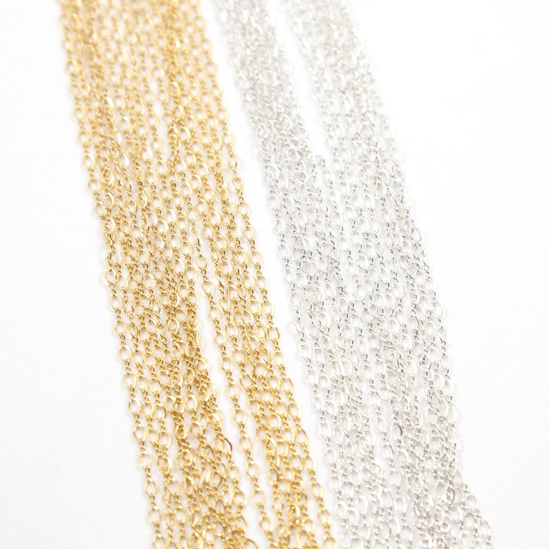 May include: Two strands of chain, one gold and one silver, are laid out side by side on a white background. The chains are both thin and delicate, and they are made of small, linked circles.