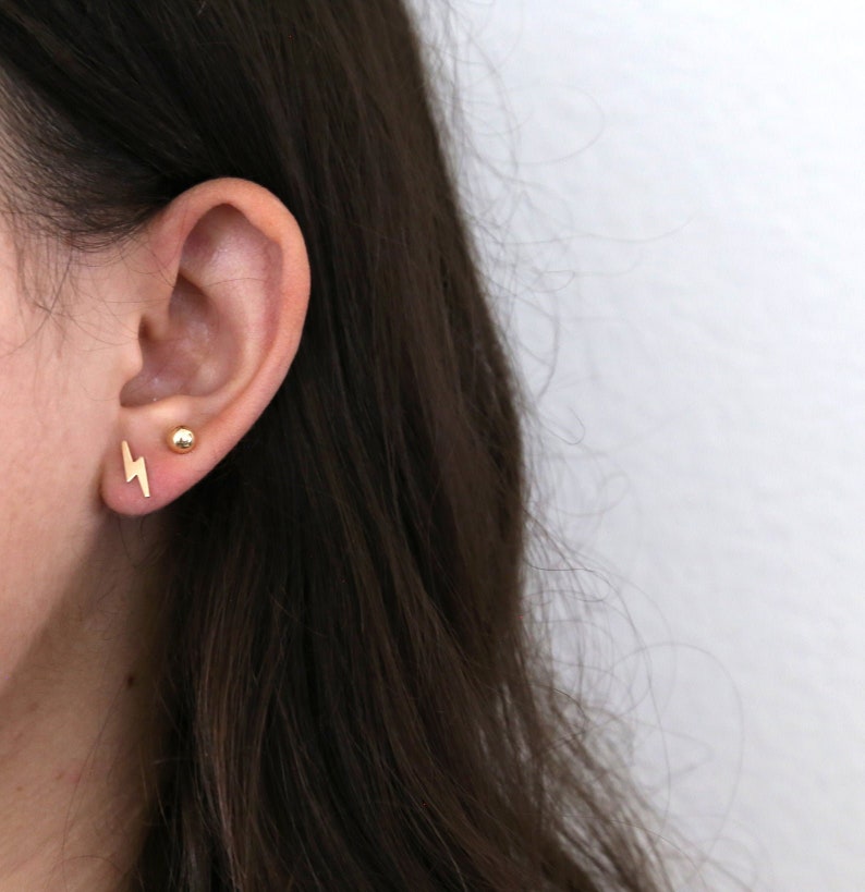 May include: A close-up of a person's ear with two gold earrings. One earring is a small, round ball and the other is a lightning bolt.