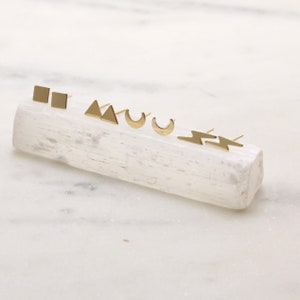 May include: A set of five gold geometric stud earrings displayed on a white crystal. The earrings are in the shapes of a square, triangle, crescent moon, and lightning bolt.