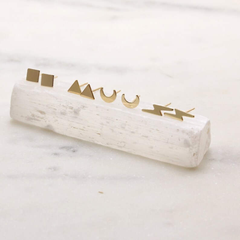 May include: A set of five gold geometric stud earrings displayed on a white crystal. The earrings are in the shapes of a square, triangle, crescent moon, and lightning bolt.