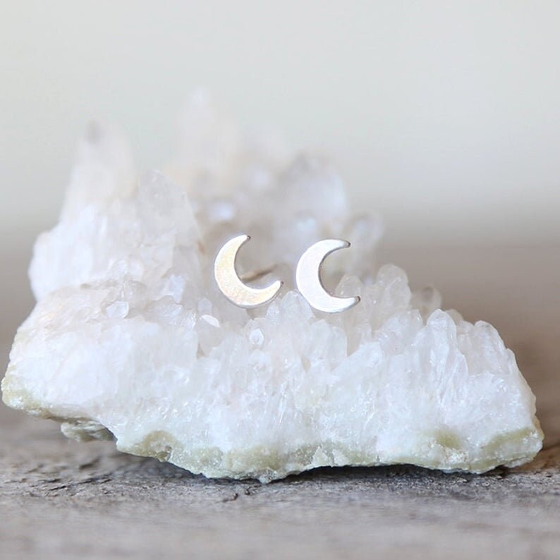 May include: A pair of silver crescent moon stud earrings on a white crystal background.