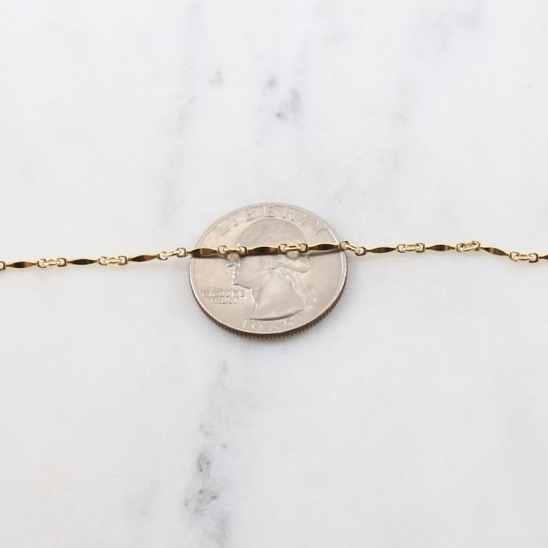 May include: A gold chain necklace with a delicate, flat, oval-shaped link. The chain is laid out on a white surface next to a US quarter for size comparison.