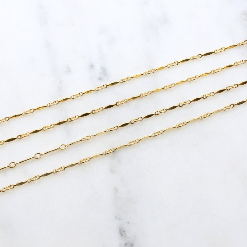 May include: Four gold chains with a delicate, flat, oval link design. The chains are layered on a white marble surface.