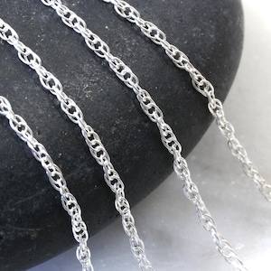 May include: Four silver chains of varying thicknesses, lying on a gray stone surface. The chains are made of a twisted rope design.