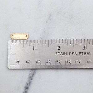 May include: A gold-colored metal tag with a textured surface. The tag is approximately 1 inch long and 1/2 inch wide. It is shown next to a ruler for size reference.