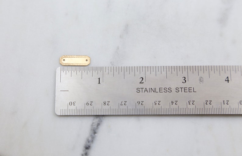 May include: A gold-colored metal tag with a textured surface. The tag is approximately 1 inch long and 1/2 inch wide. It is shown next to a ruler for size reference.