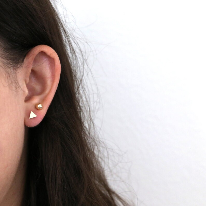 May include: A close-up of a person's ear with two gold earrings. One earring is a small, round ball and the other is a small, triangular shape.