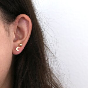 May include: A close-up of a person's ear with two gold earrings. One earring is a small, round ball and the other is a crescent moon.