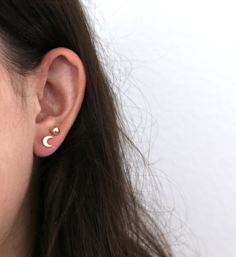 May include: A close-up of a person's ear with two gold earrings. One earring is a small, round ball and the other is a crescent moon.